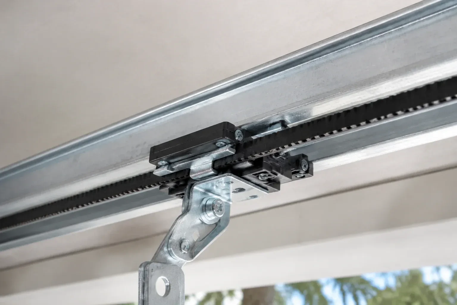 Black plastic trolley and bent galvanized bracket on a steel opener rail with a toothed black belt, against a ceiling. — Belt-Drive Opener Repair, Kawartha Lakes