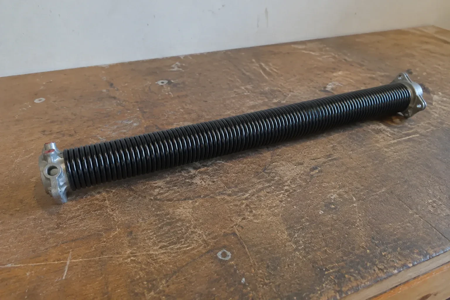 Detached black coil spring with metal end fittings on a workbench. — Torsion Spring Replacement, Kawartha Lakes