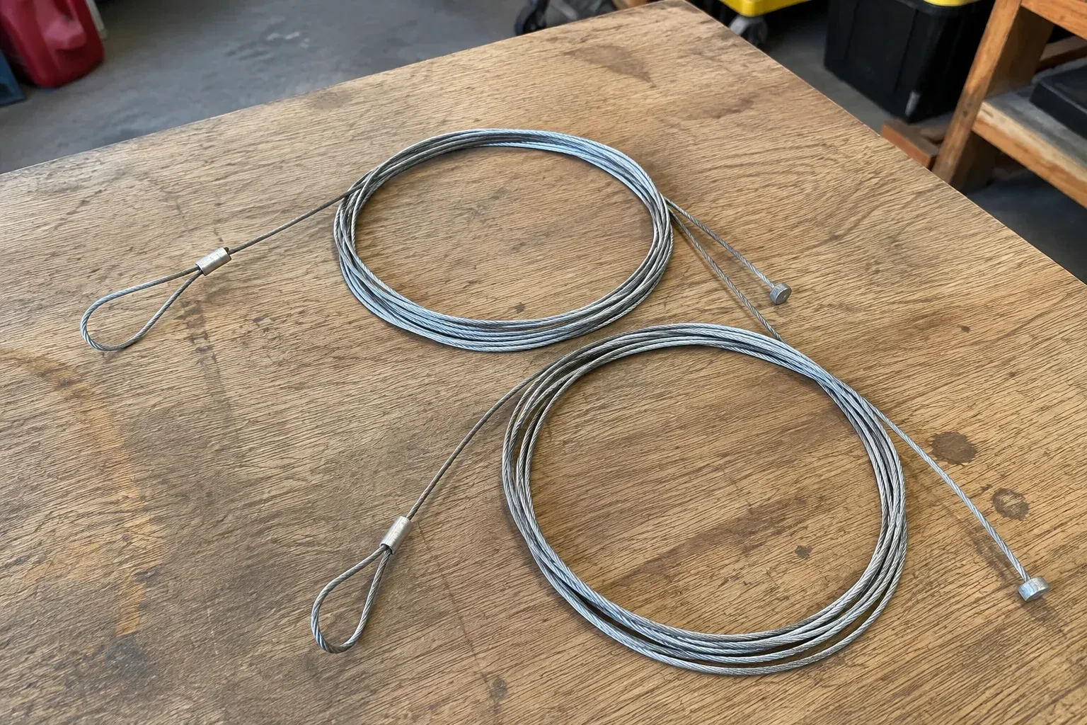 Two coiled steel cables with loop and stop ends on a workbench. — Garage Door Cable Replacement, Kawartha Lakes